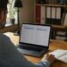 Mezczyzna przy biurku porownuje na laptopie dwie tabele ofert ubezpieczenia, obok notatnik z wyliczeniami